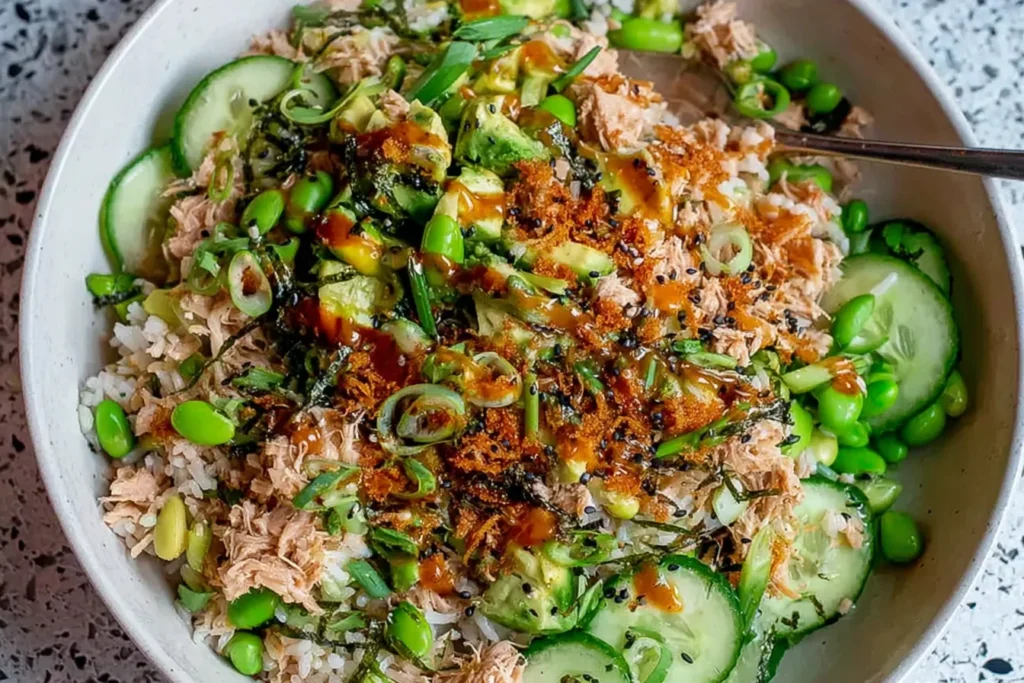 Tuna Avocado Crispy Rice Salad bowl with flaky tuna, cucumber slices, edamame, avocado, scallions, sesame seeds, and spicy drizzle over rice.