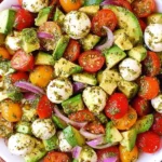 Avocado Salad Tomatoes Mozzarella Basil Pesto in a white bowl with red and yellow cherry tomatoes, avocado chunks, cucumber slices, red onion, and mozzarella pearls.
