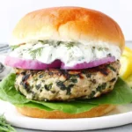 Chicken Burgers with Feta Cheese & Spinach on a brioche bun with tzatziki, red onion, and lettuce on a plate with cucumber slices.