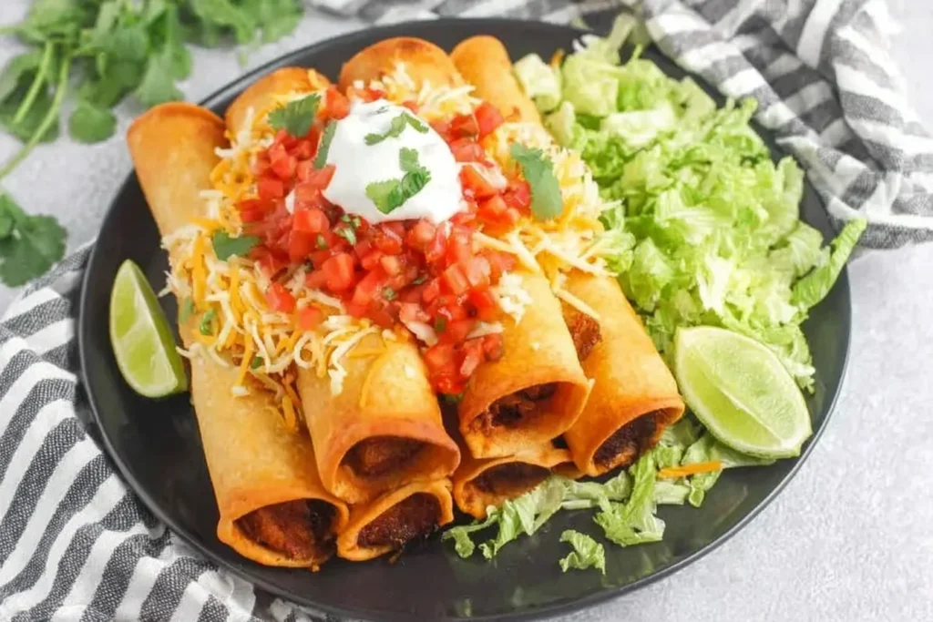 Plate of crispy Chicken Flautas topped with shredded cheese, pico de gallo, sour cream, and cilantro, served with lettuce and lime wedges.