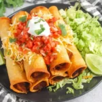 Plate of crispy Chicken Flautas topped with shredded cheese, pico de gallo, sour cream, and cilantro, served with lettuce and lime wedges.