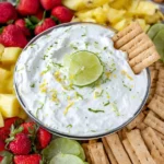 Key Lime Pie Fruit Dip in a glass bowl topped with lime zest and a lime slice, served with strawberries, pineapple chunks, and graham crackers.