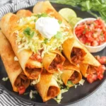 Crispy Chicken Flautas topped with shredded cheese, lettuce, pico de gallo, and sour cream on a black plate with lime wedges.