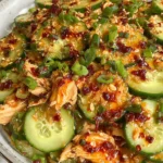 Thai Cucumber Salad with Chicken in a bowl with sliced cucumbers, shredded chicken, peanut-lime dressing, green onions, sesame seeds, and chili crisp