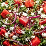 Feta Watermelon Salad with arugula, red onion, mint, and crumbled feta in a balsamic honey dressing, served in a black bowl.