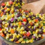 Corn and Black Bean Salad with Mexican Vinaigrette in a glass bowl, mixed with red onion, bell peppers, and cilantro, with a wooden spoon.