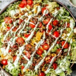Taco Salad topped with seasoned ground beef, romaine lettuce, cherry tomatoes, crunchy tortilla chips, and creamy cilantro-lime dressing drizzle.