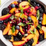 Honey Lime Basil Peach Fruit Salad with sliced peaches, blackberries, blueberries, pomegranate seeds, and fresh basil in a bowl with lime.