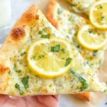 Hand holding a slice of Lemon Pizza with melty mozzarella, lemon slice, and fresh herbs—zesty white pizza perfect for summer.