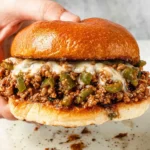 Hand holding a Philly Cheesesteak Sloppy Joes sandwich on a toasted brioche bun, packed with saucy beef, green peppers, and melted provolone.