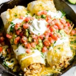 Smothered Honey Lime Chicken Burritos smothered in creamy salsa verde, melted cheese, pico de gallo, and sour cream in a cast-iron skillet.