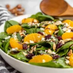Spinach Mandarin Salad in a white bowl with baby spinach, mandarin orange slices, dried cranberries, toasted almonds, goat cheese, and a honey balsamic drizzle.