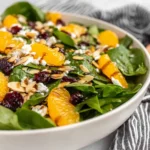 Spinach Mandarin Salad in a white bowl with baby spinach, mandarin oranges, dried cranberries, toasted sliced almonds, and goat cheese crumbles.
