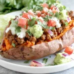 Taco Stuffed Sweet Potatoes on a plate with seasoned ground beef, guacamole, pico de gallo, cilantro, and sour cream, served with lime wedges.