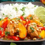 Thai Peach Chicken served over fluffy white rice with peach slices, red bell pepper, chopped peanuts, fresh herbs, and lime wedges.
