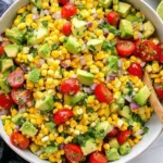 Summer Corn Salad with Avocado in a large bowl with cherry tomatoes, red onion, cilantro, and fresh lime wedges on the side.