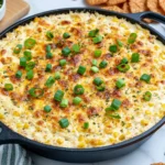 Baked Elote Dip in a cast-iron skillet topped with melted golden cheese, sweet corn, and fresh green onions, served with crackers for dipping.