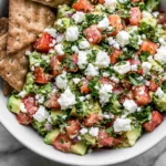 Avocado Feta Dip with ripe avocado, feta cheese, tomatoes, red onion, parsley, and crackers served in a bowl.