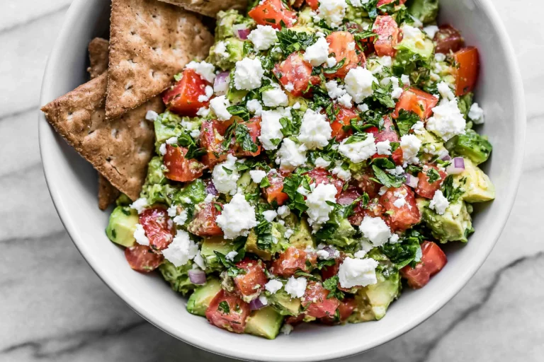 Avocado Feta Dip with ripe avocado, feta cheese, tomatoes, red onion, parsley, and crackers served in a bowl.