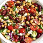 Mediterranean Dense Bean Salad with chickpeas, kidney beans, cucumbers, cherry tomatoes, red onion, feta, and herbs in a white bowl.