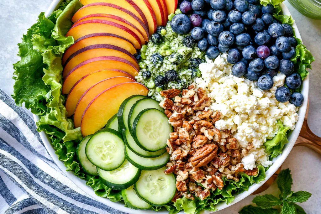 Peach Berry Summer Salad featuring sliced peaches, fresh blueberries, cucumber rounds, feta cheese, chopped pecans, and leafy greens with lemon basil dressing.