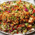 Sesame Chicken Cabbage Crunch Salad on a platter with sliced sesame chicken, shredded cabbage, carrots, cucumbers, peppers, herbs, and sesame ginger dressing.